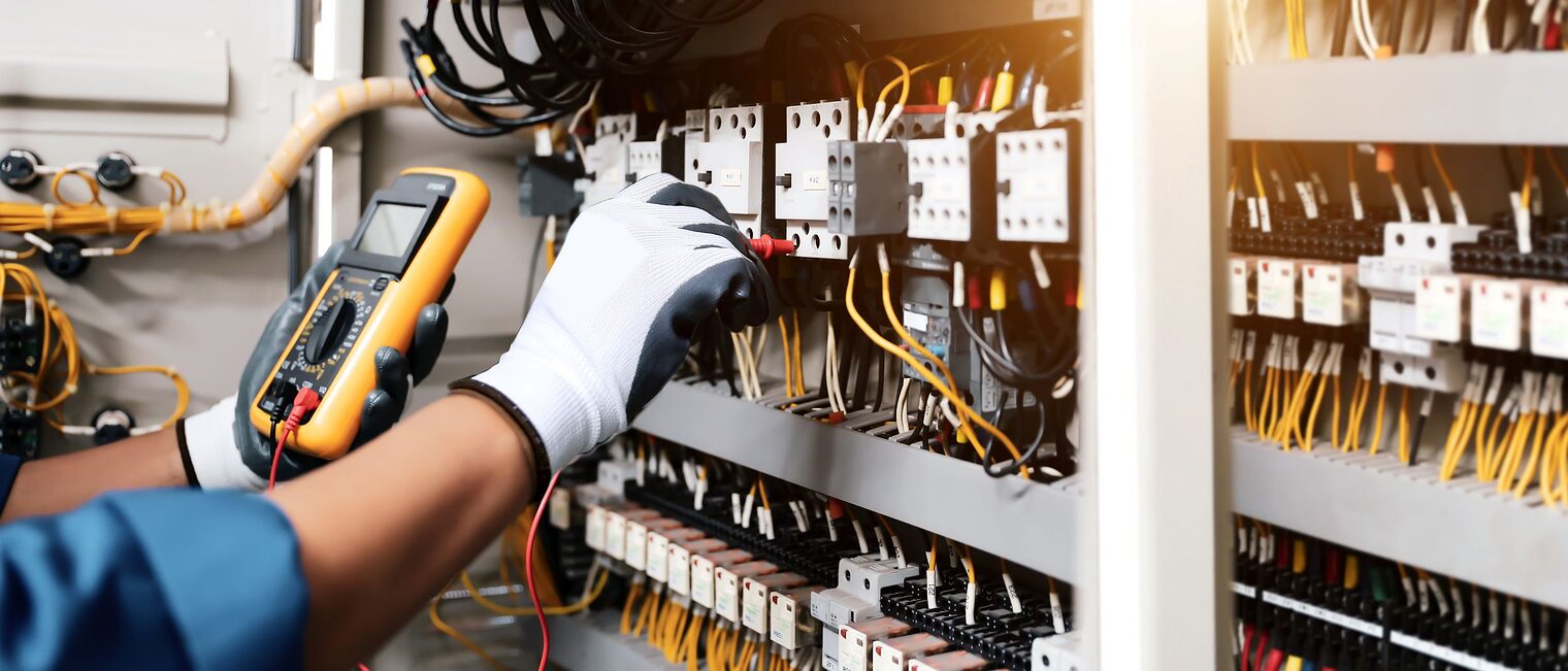Elektriker testet elektrische Verbindungen in einer Schalttafel mit Multimeter. Nahaufnahme eines Schaltschrankes mit Kabeln, Schaltern und Sicherungen. Eine Person mit Handschuhen misst elektrische Spannungen mit einem digitalen Multimeter.